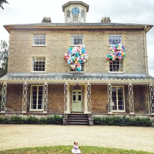 Victorian-style house with colorful balloons in windows, clock tower, and child sitting on the lawn.
