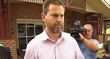 A man in a light-colored shirt walking outside a building as a cameraman films him from behind.