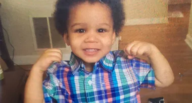 A toddler in a colorful plaid shirt smiling and holding up both fists indoors.