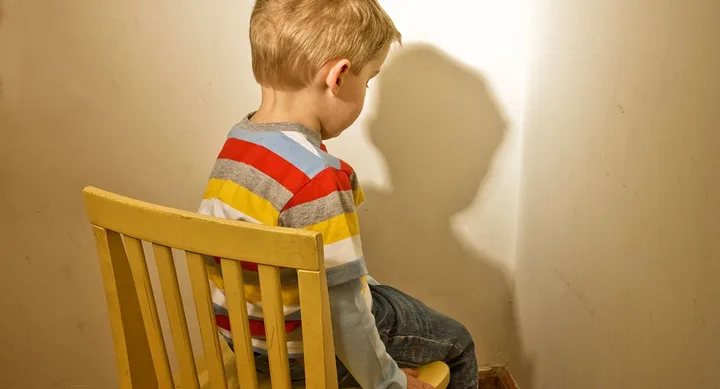 Child in a striped shirt sitting on a yellow chair facing the corner of a dimly lit room.