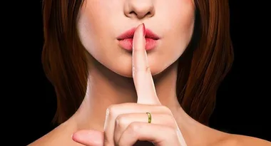 Woman with index finger on lips, suggesting silence, against a black background.