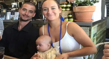 A couple smiling, seated at a cafe, holding a baby in their arms, with plants and shelves in the background.
