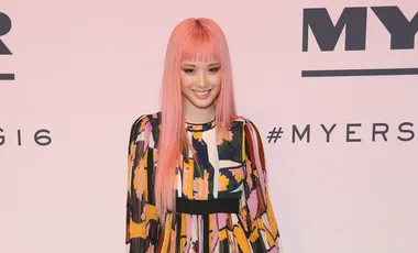 Young woman with pink hair in a colorful patterned dress, smiling at a fashion event with a branded backdrop.