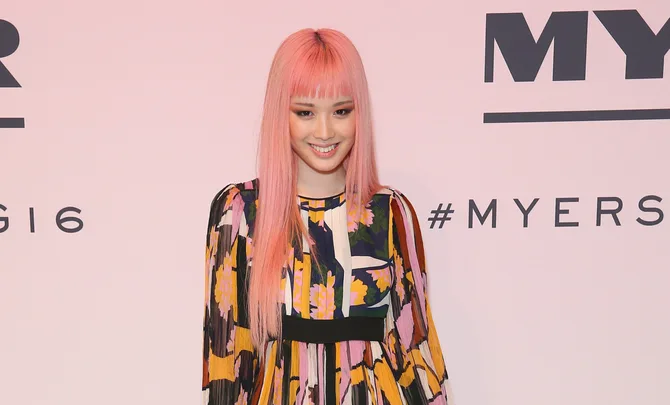 Young woman with pink hair in a colorful patterned dress, smiling at a fashion event with a branded backdrop.