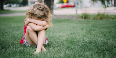 A young child in a red top sits on grass with head resting on crossed arms, appearing thoughtful or upset.