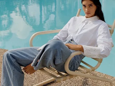 A woman in a white shirt and jeans sits on a lounge chair by a pool, with a calm expression and her feet bare.