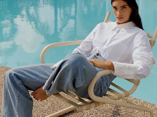 A woman in a white shirt and jeans sits on a lounge chair by a pool, with a calm expression and her feet bare.