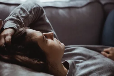 A woman lying on a couch with her arm resting on her forehead, looking contemplative.