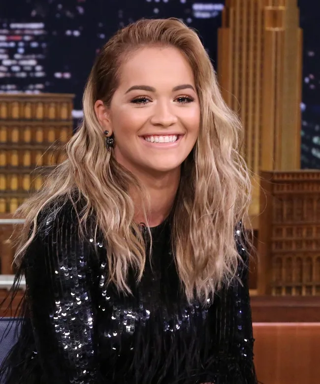 Person smiling with long, wavy blonde hair, wearing a black sequined top, against a cityscape background.