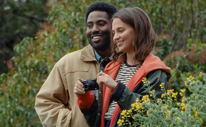 Two people standing outside, smiling and holding a camera, surrounded by greenery and yellow flowers.