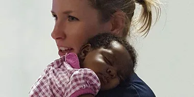 Woman holding a sleeping baby in a pink checkered outfit against her shoulder.