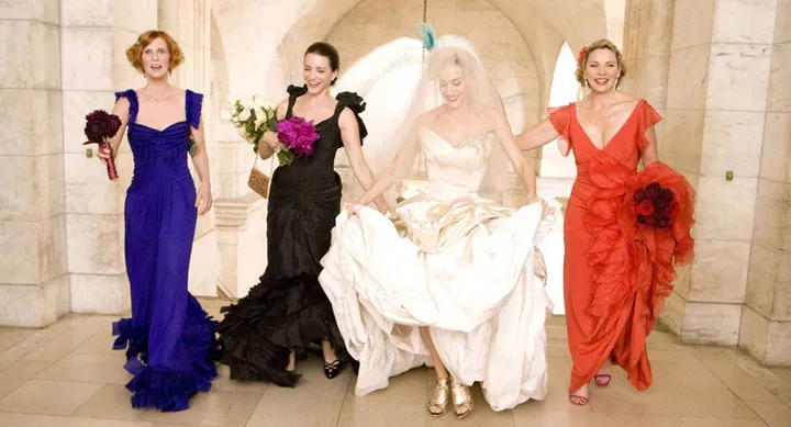 Four women in colorful dresses, including a bride, walking joyfully in a grand hallway.