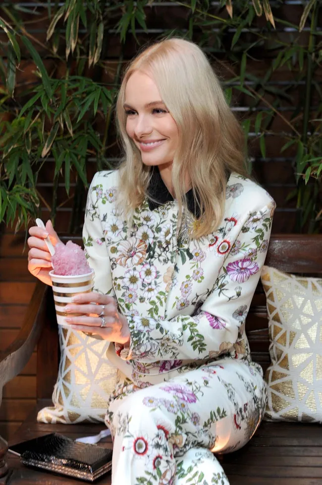 Blonde woman in floral outfit smiling, holding a cup with pink shaved ice, seated on a wooden bench with greenery background.
