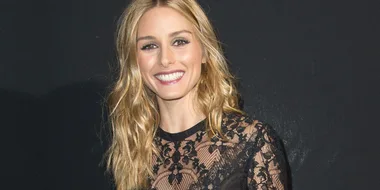A person with long blonde hair smiles, wearing a lace black top against a dark background.