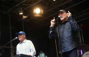 Two men wearing hats perform on stage, one singing and playing keyboard, the other gesturing with both hands.