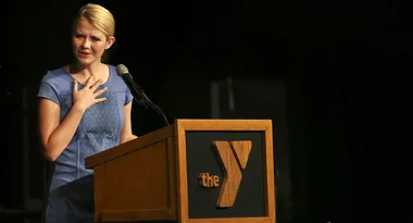 A woman speaks at a podium with a YMCA logo, wearing a blue dress and holding a microphone.