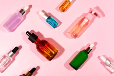 Various colored cosmetic serum bottles arranged on a pink background, some with droppers, others with pumps.