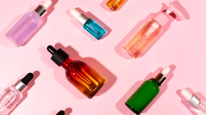 Various colored cosmetic serum bottles arranged on a pink background, some with droppers, others with pumps.