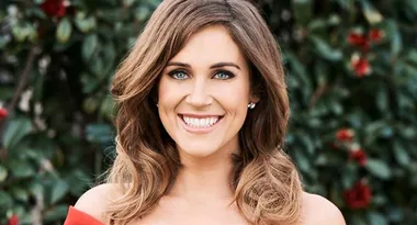 A woman with wavy brown hair smiles at the camera, standing in front of a leafy background.