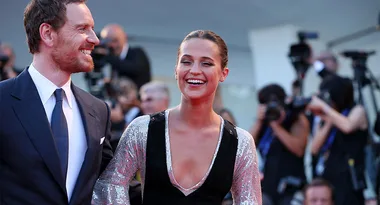 Two people smiling and walking on a red carpet, surrounded by photographers.