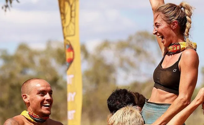 Group celebrating outdoors, woman lifted with arm raised, wearing a black top and buff.