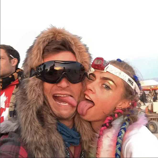 Two people at a festival pose playfully with tongues out, wearing furry accessories and goggles.