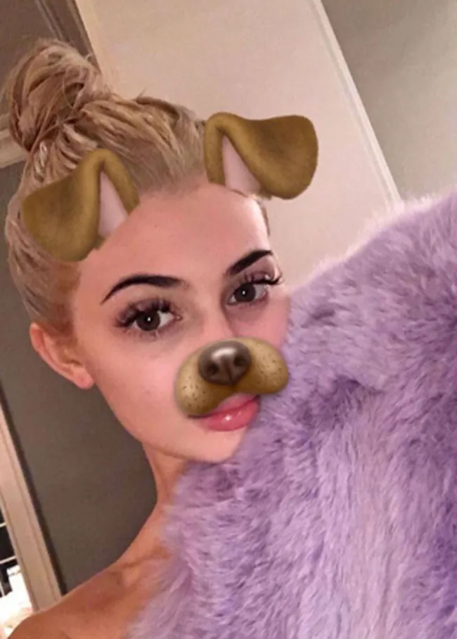Young woman with a dog face filter, blonde hair in a bun, holding a purple furry item.