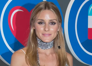 A woman with long hair, wearing a patterned choker and dangling earring, stands in front of a colorful heart background.