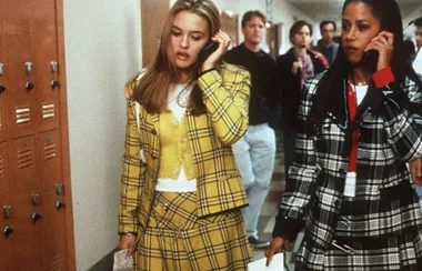 Two women in plaid outfits walking in a school hallway, talking on mobile phones.