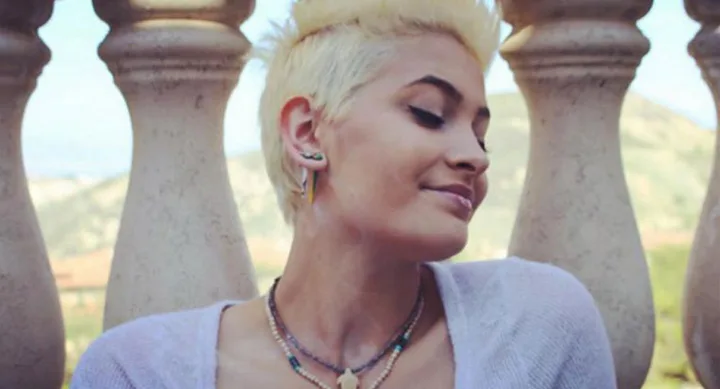 A person with short blonde hair smiles with eyes closed, standing against a stone balustrade.