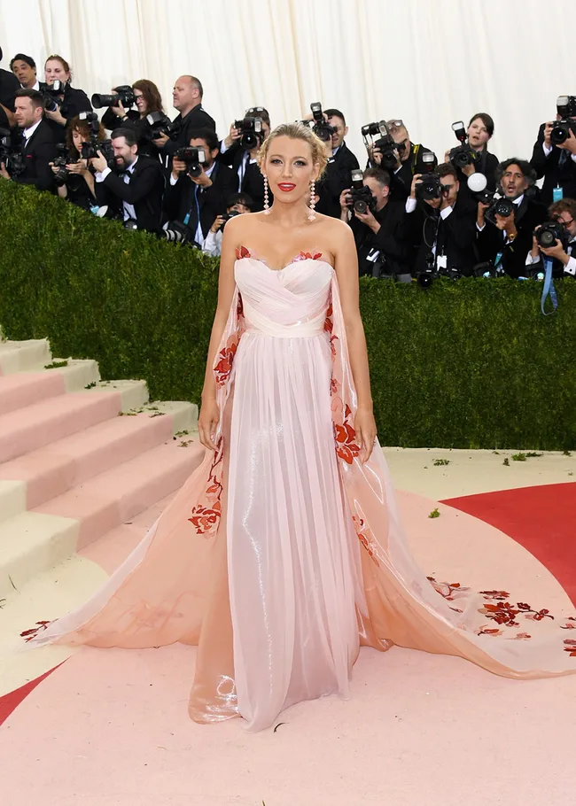Blonde woman in a floral pink gown poses on a red carpet, surrounded by photographers.