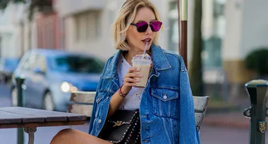 Woman in a denim jacket and sunglasses drinks iced coffee at an outdoor cafe table.