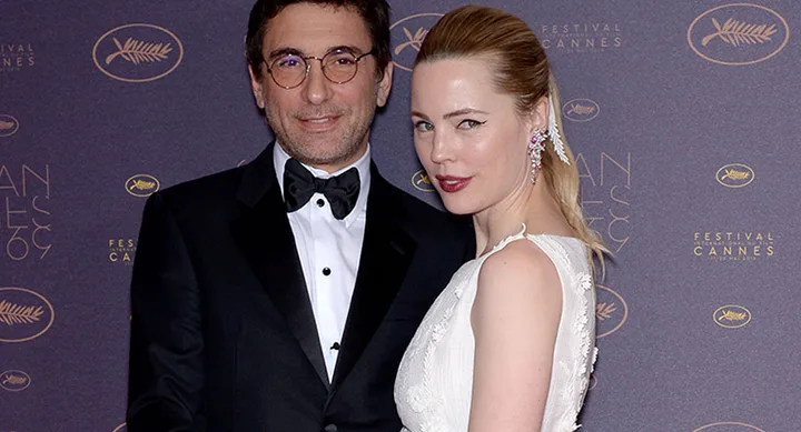Man in a tuxedo and woman in a white dress pose on the Cannes Film Festival red carpet.