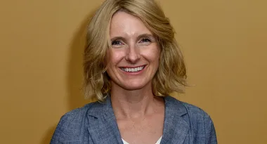 A person with shoulder-length blonde hair, wearing a blue blazer, smiling in front of a beige background.