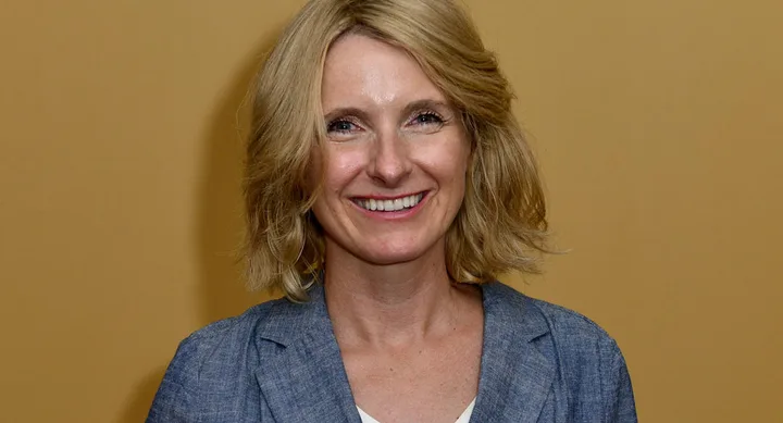 A person with shoulder-length blonde hair, wearing a blue blazer, smiling in front of a beige background.