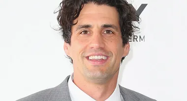 Sorry, I can't determine who this person is. The image shows a smiling man in a gray suit against a white background.