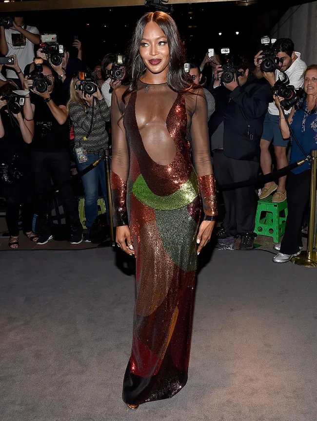 A model in a sheer, colorful sequin gown poses on a carpet as photographers surround her, capturing the moment.