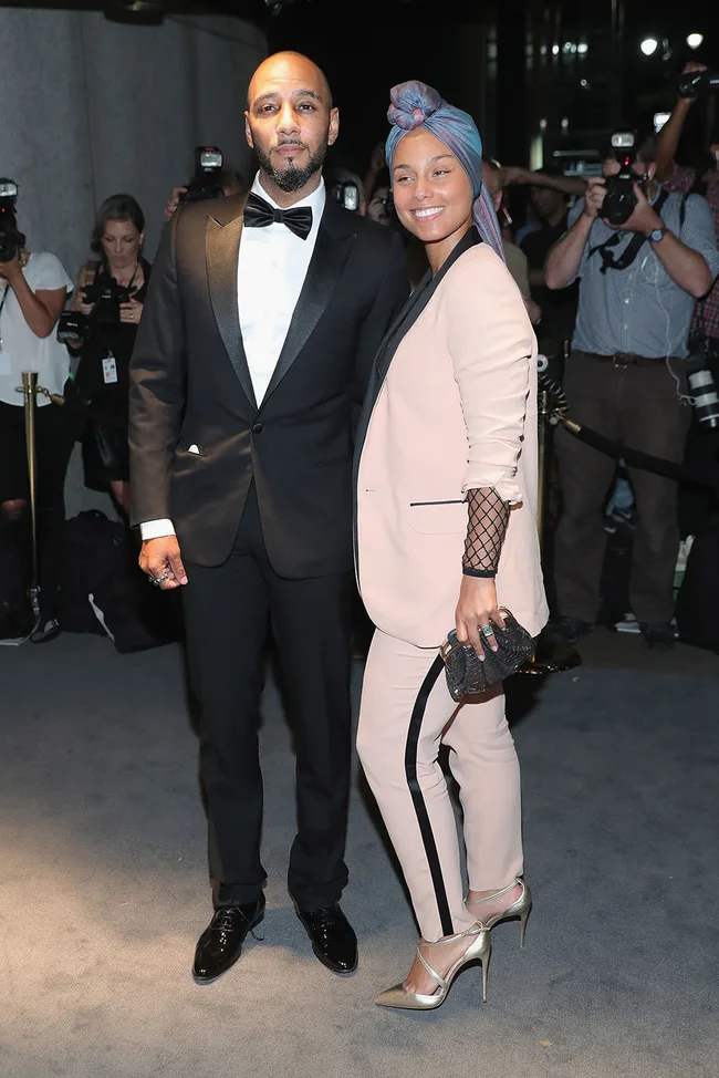 A man in a tuxedo and a woman in a pink suit pose on a red carpet, photographers in the background.