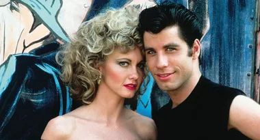 Two actors, one with curly blonde hair and the other with dark hair, stand closely together against a painted wall.