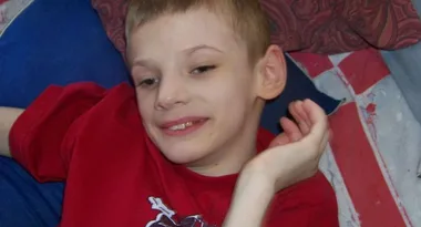 Boy in a red shirt smiling, resting on a couch with a patterned background.