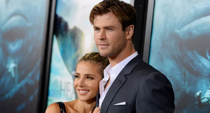 Man in a suit and woman in a dress smiling at a movie premiere with a poster backdrop.