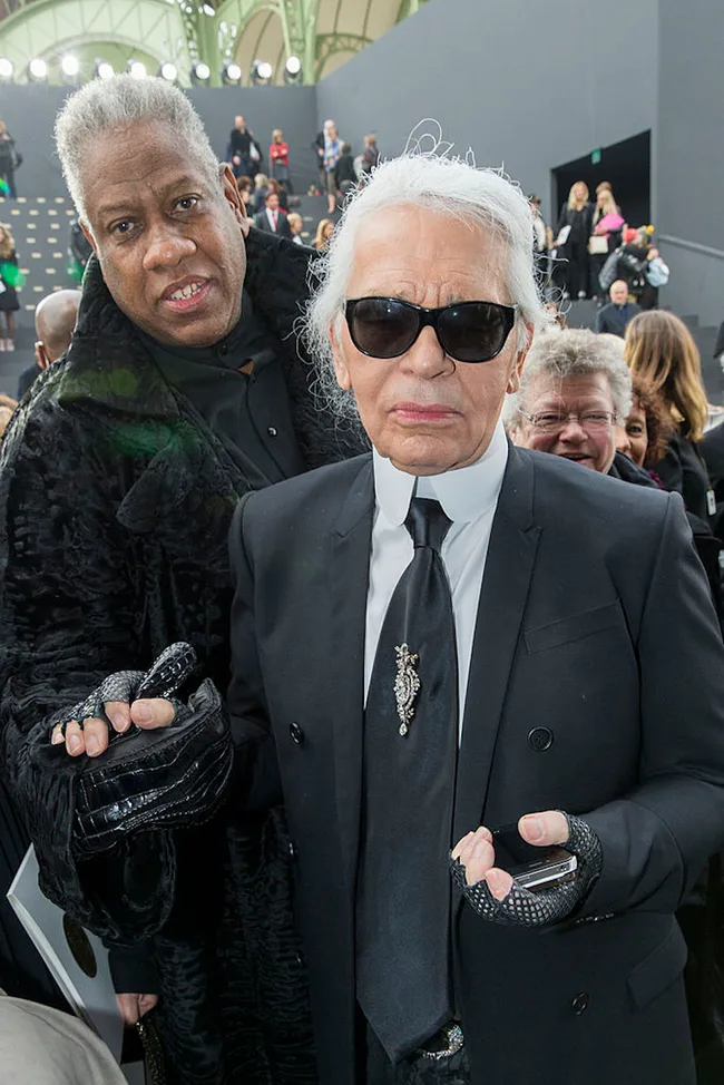 Two stylish individuals in formal attire, one with sunglasses, attending a fashion event, holding hands.