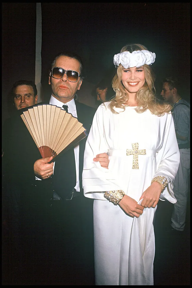 Two people pose at an event; one in a suit with sunglasses and fan, the other in a white gown with floral headpiece.