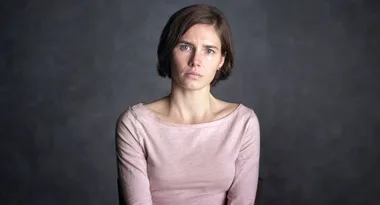 A person with short hair in a light pink top, looking serious against a dark background.
