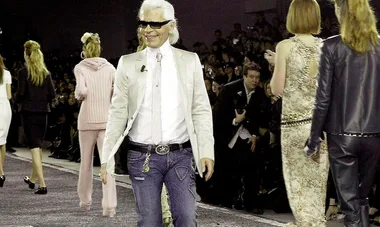 Designer wearing white jacket and sunglasses walking on a fashion runway, surrounded by models, with audience in background.