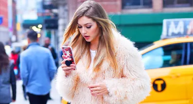 A woman in a fuzzy coat looks at her phone, walking on a city street with a taxi in the background.