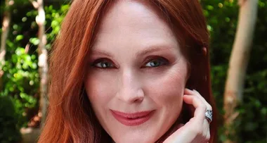 A woman with long red hair and a gentle smile, touching her chin with her hand, posing outdoors with greenery in the background.