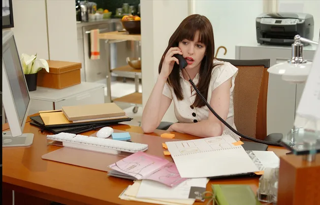 A woman with dark hair sits at a cluttered desk, talking on a corded phone, surrounded by office items and a computer.