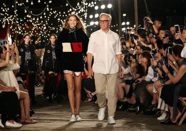 Model and designer walking on a lit runway with an audience taking photos.
