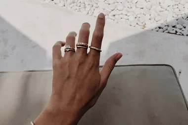 A hand with multiple rings, resting on a cushioned surface with white pebbles in the background.
